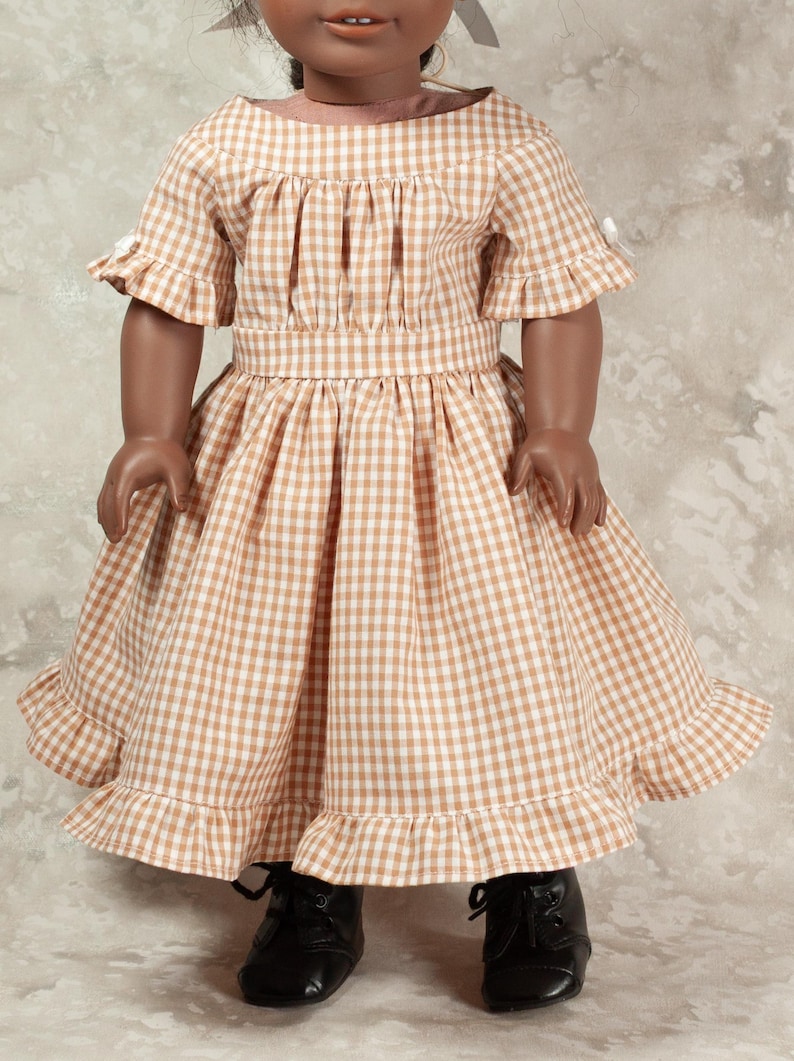 May include: A doll wearing a tan and white gingham dress with a gathered bodice, short ruffled sleeves, and a ruffled hemline. The dress features a fitted waistband and is paired with black lace-up shoes. The doll has dark skin and dark hair.