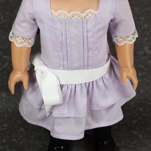 May include: A lavender doll dress with a square neckline and short sleeves trimmed with white lace. The dress features a white ribbon belt and a layered skirt. The doll is wearing black shoes.