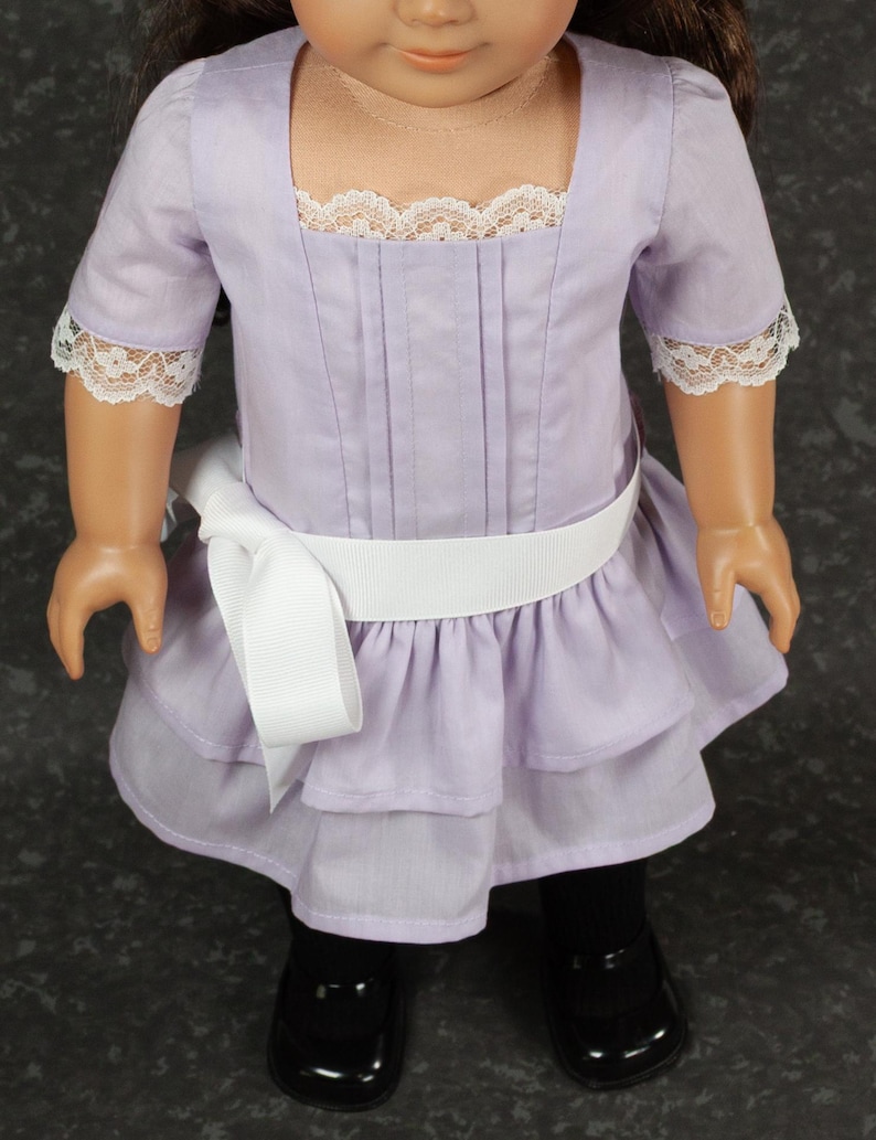 May include: A lavender doll dress with a square neckline and short sleeves trimmed with white lace. The dress features a white ribbon belt and a layered skirt. The doll is wearing black shoes.
