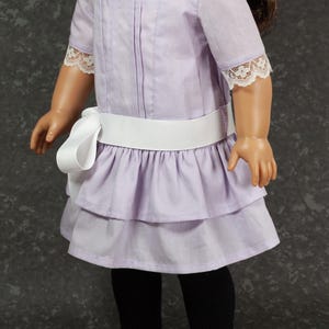 May include: A doll wearing a lavender dress with a white ribbon belt and lace trim. The dress has a tiered skirt and short sleeves. The doll is also wearing black tights and black shoes.
