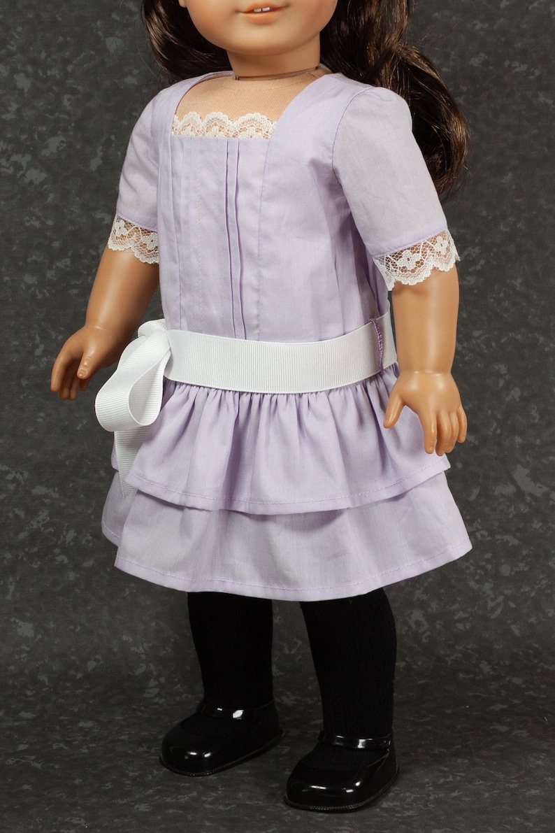 May include: A doll wearing a lavender dress with a white ribbon belt and lace trim. The dress has a tiered skirt and short sleeves. The doll is also wearing black tights and black shoes.