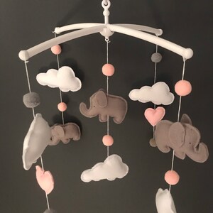 May include: A baby mobile with grey and pink felt elephants, clouds, and hearts hanging from a white metal frame. The mobile is decorated with pink and grey pom-poms.
