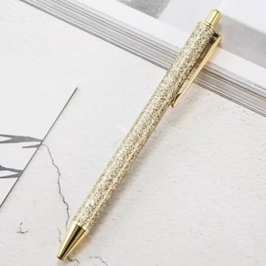 Può includere: Una penna dorata con glitter e un fermaglio in metallo dorato. La penna ha una punta a sfera.