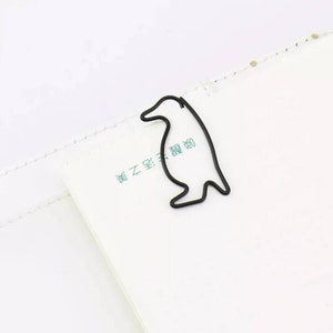 Op de afbeelding: Een zwarte metalen paperclip in de vorm van een pinguïn. De clip bevindt zich op een wit oppervlak met de tekst "書之星" in groen.