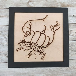 May include: A wood framed sign with a laser-cut design of a large hand with three smaller hands holding onto it. The large hand has the word "Dad" written on it. The smaller hands are labeled "Genevieve", "Colton", and "Damian".