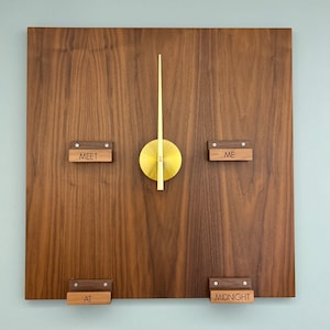 May include: A square wooden wall clock with a dark brown finish. The clock features a gold-colored circular center with gold hands. The words "MEET", "ME", "AT", and "MIDNIGHT" are displayed on small wooden blocks.
