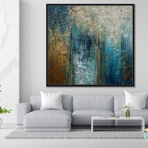May include: Abstract artwork in a black frame. The painting has a textured surface with vertical strokes in blue, gold, and brown. It hangs above a grey sofa with white cushions, a coffee table, and a small potted plant. The artwork is a modern piece.
