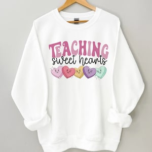 以下が含まれることがあります： ピンクとパープルの文字で「TEACHING sweet hearts」と書かれた白いスウェットシャツ。テキストの下には、顔が付いたカラフルなキャンディーハートが6つ並んでいます。
