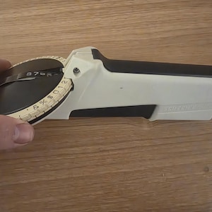 May include: A handheld manual embossing label maker with a cream-colored plastic body and a black circular dial. The dial features embossed letters, numbers, and symbols including an ampersand and dollar sign. The device has a black handle grip.