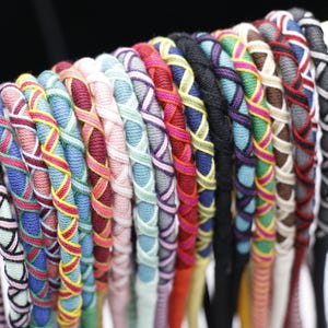 May include: A collection of colorful friendship bracelets, each with a unique woven design. The bracelets feature a variety of colors, including blue, pink, green, and black, and are displayed on a wooden dowel. The intricate patterns add a handmade touch.