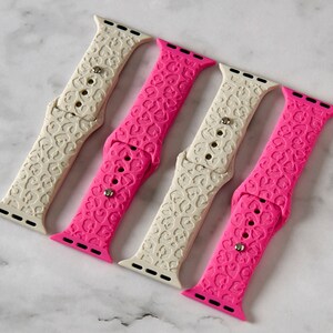 May include: Four Apple Watch bands in pink and white with a leopard print design. The bands are made of silicone and have a heart pattern.