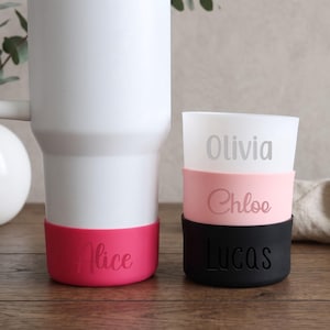 May include: A white tumbler with a hot pink silicone boot personalised with the name Alice. Next to it, a stack of three silicone boots in white, light pink, and black, personalised with the names Olivia, Chloe, and Lucas respectively.