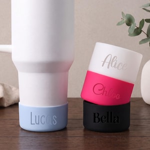 May include: A white tumbler with a light blue silicone boot featuring the name Lucas. Beside it, a stack of three silicone boots in white, hot pink, and black, personalised with the names Alice, Chloe, and Bella respectively in script font.