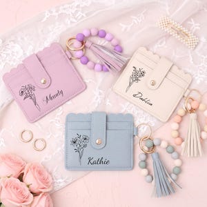 May include: Three rectangular card holders with scalloped edges and snap closures in lavender, cream, and light blue. Each features a floral line drawing and a name: Mandy, Dahlia, or Kathie. Each includes a silicone bead wristlet and tassel.