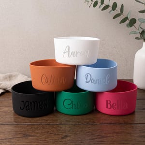 May include: A stack of six colourful silicone protective boots for tumblers, each featuring a unique engraved name. The colours include white with Aaron, orange with Calvin, light blue with Daniel, black with James, green with Chloe, and pink with Bella.