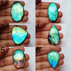 Può includere: Una collezione di sei pietre preziose di labradorite lucidata in varie forme. Le pietre mostrano lampi iridescenti di blu, verde e giallo. Ogni pietra è tenuta in una mano, mostrando la bellezza naturale e i motivi unici.