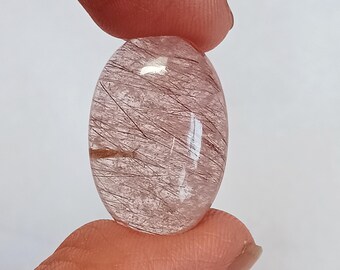 May include: Oval-shaped gemstone with a translucent, pinkish hue. Fine, dark, needle-like inclusions are visible throughout the stone. The gemstone is held between two fingers, showcasing its polished surface and natural beauty.