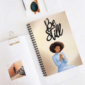 May include: A spiral-bound notebook featuring a beige cover with the black text "Be Still" and an illustration of a person with dark curly hair wearing a light blue long-sleeved dress with hands clasped together in a prayer-like position.