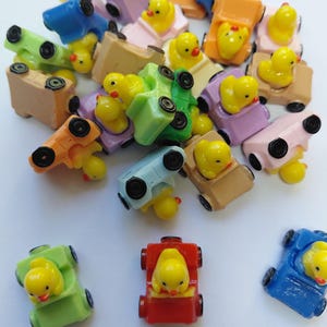 May include: A collection of small plastic figurines featuring yellow ducks sitting in miniature cars of various colours including red, blue, green, orange, pink, and tan. Each car has four black wheels and the ducks have orange beaks and black eyes.