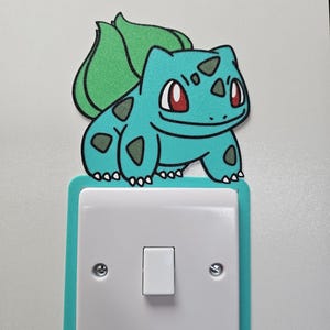 May include: A light switch cover plate featuring a teal and green Bulbasaur character graphic positioned above a white square light switch. The decorative plate has a teal border and is mounted on a flat surface, showcasing the creature's design.