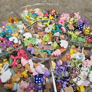May include: A clear, segmented tray overflowing with colourful charms and keyrings. The charms feature ice cream cones, bears, dice, and balloon animals. Keyrings with silver rings are attached to many of the charms. The tray is on a neutral surface.