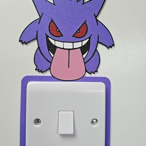 May include: A purple Gengar character decal positioned above a white square light switch. The character features red eyes, a wide grin with white teeth, and a long pink tongue hanging down. A purple border frame surrounds the light switch.