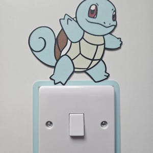 May include: A light blue light switch surround featuring a cartoon turtle character with a brown shell and cream-coloured belly. The character is positioned above a white rectangular light switch plate with two visible silver screws on a flat surface.