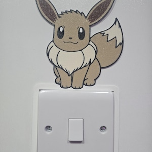 May include: A light switch cover featuring a decorative sticker of the brown and cream-coloured Eevee character from the Pokémon franchise. The sticker is positioned directly above a standard white square light switch with two visible screws.