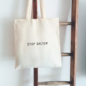 Könnte beinhalten: Eine weiße Stofftasche mit dem Aufdruck "STOP RACISM" in Schwarz auf der Vorderseite. Die Tasche hängt an einer Holzleiter.
