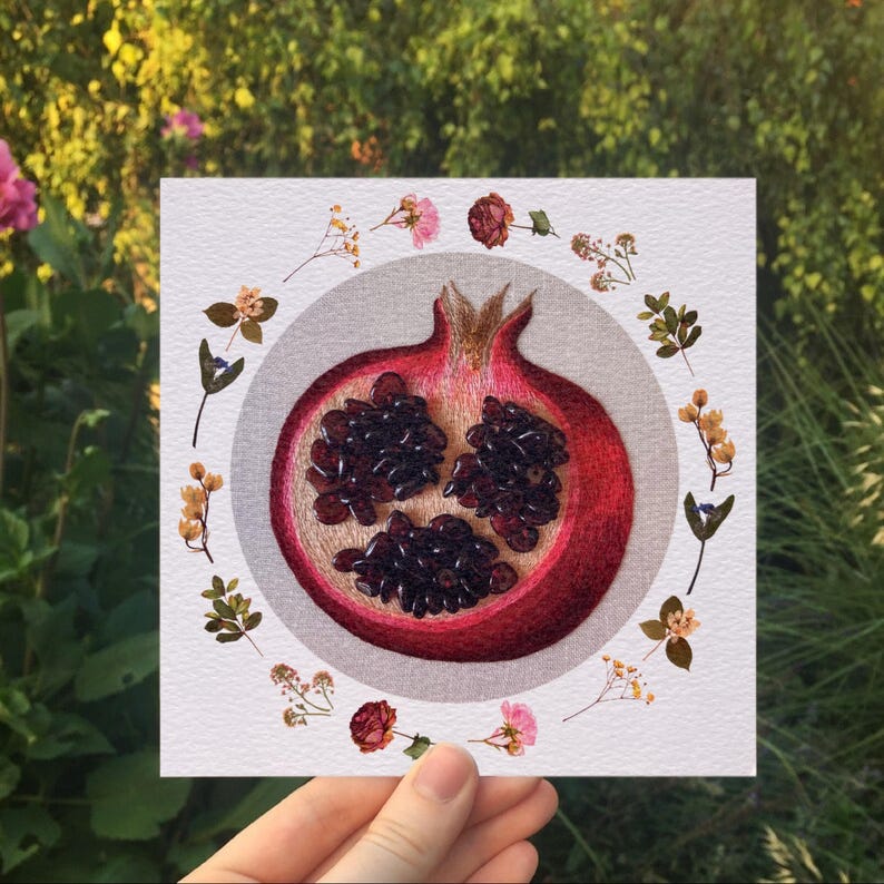 May include: A close-up of an embroidered artwork depicting a red pomegranate cut open, revealing the seeds inside. The pomegranate is surrounded by a circle of dried flowers and leaves.
