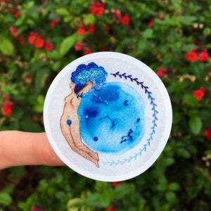 Op de afbeelding: Een witte ronde sticker met een blauw en wit geborduurd ontwerp van een vrouw met blauw haar die een blauwe maan met sterren en sterrenbeelden vasthoudt.