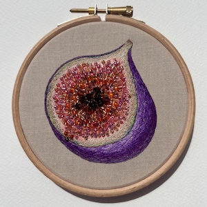 May include: Embroidered fig art in a wooden hoop. The fig is depicted in a cross-section, showcasing a vibrant interior of pink, orange, and brown beads. The outer skin is a deep purple, with green and tan accents. The embroidery is set against a neutral fabric.