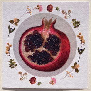 May include: A red pomegranate with seeds visible inside, surrounded by a circle of dried flowers on a white background.