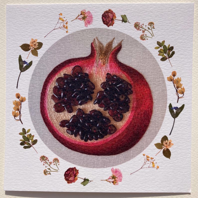 May include: A red pomegranate with seeds visible inside, surrounded by a circle of dried flowers on a white background.