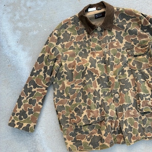 May include: A long-sleeve jacket featuring a multi-colored camouflage pattern in shades of brown, green, and tan. The jacket includes a dark brown corduroy collar, a button-front closure, two large lower front pockets, and a chest pocket.