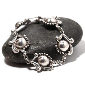 May include: A silver bracelet with a series of round, polished beads and decorative, curved links. The bracelet is displayed on a dark gray, textured stone, showcasing its intricate design and metallic sheen. The bracelet's clasp is visible.