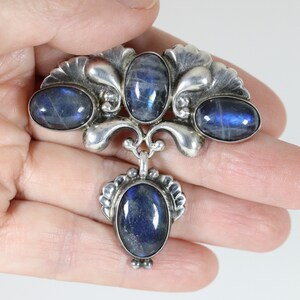 Puede incluir: Un broche de plata con cuatro piedras ovaladas de labradorita azul engastadas en un dise&ntilde;o floral. El broche tiene un colgante con una quinta piedra.
