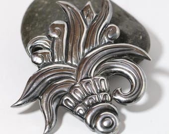 Könnte beinhalten: Eine silberfarbene, verzierte Metallbrosche mit einem stilisierten floralen Design. Die Brosche hat geschwungene Linien, Wirbel und spitze Elemente. Sie liegt auf einem glatten, grauen Stein vor weißem Hintergrund.