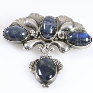 Puede incluir: Un broche de plata con cuatro piedras azules ovaladas engastadas en un dise&ntilde;o decorativo. El broche tiene un colgante con una piedra azul.
