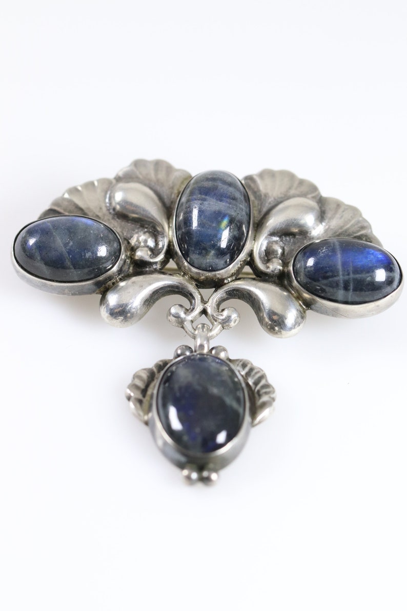 Puede incluir: Un broche de plata con cuatro piedras azules ovaladas engastadas en un dise&ntilde;o decorativo. El broche tiene un colgante con una piedra azul.