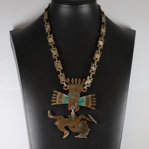 May include: A gold and copper necklace with a large pendant featuring a stylised figure with wings and a crown. The pendant is decorated with turquoise and other colourful enamel.