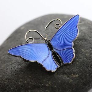 May include: A blue butterfly brooch with silver-colored accents. The wings are a vibrant blue with detailed lines, and the body is black. The brooch is set against a dark gray background.