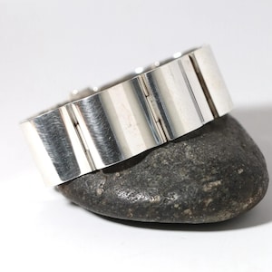 May include: A silver-toned bracelet with a rectangular design. The bracelet is composed of several connected rectangular segments, each with vertical grooves. The bracelet is resting on a dark gray stone, showcasing its metallic sheen.