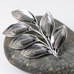 May include: A silver brooch in the shape of a stylized branch with five leaves. Each leaf is layered and textured, and each leaf has a cluster of small, round beads at the tip.
