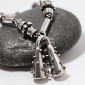 May include: A silver-toned metal bracelet featuring a chain of cylindrical and gear-shaped links. The focal point consists of two dangling, cone-shaped charms, each adorned with multiple small, protruding spikes extending from the surface.