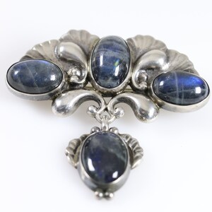 Puede incluir: Un broche de plata con cuatro piedras preciosas azules ovaladas engastadas en un dise&ntilde;o floral decorativo. El broche tiene un colgante que cuelga con una quinta piedra preciosa.
