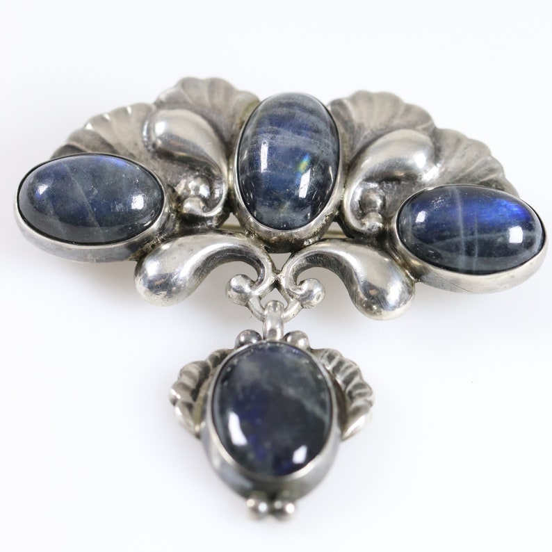 Puede incluir: Un broche de plata con cuatro piedras preciosas azules ovaladas engastadas en un dise&ntilde;o floral decorativo. El broche tiene un colgante que cuelga con una quinta piedra preciosa.