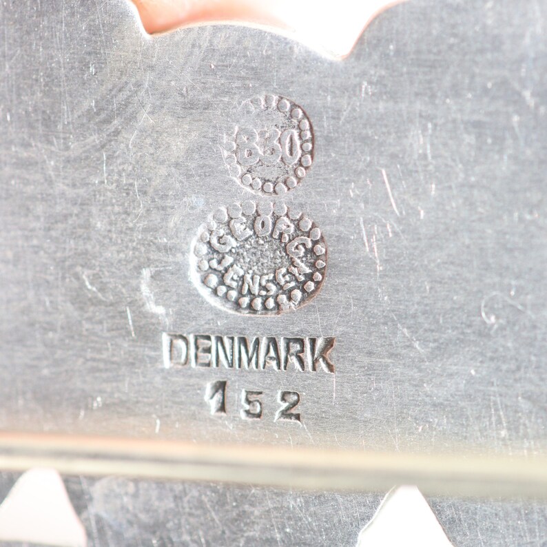 Puede incluir: Primer plano de un sello de plata con el texto "Georg Jensen" y "Denmark 152" junto con los n&uacute;meros "830" en un c&iacute;rculo.