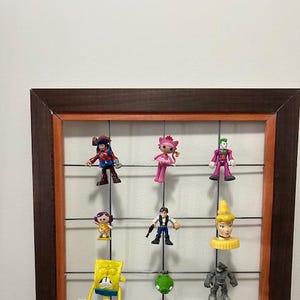 Pode incluir: Uma moldura de madeira castanho com uma grelha de arame no interior. A grelha contém várias figurinhas pequenas, incluindo um pirata, uma fada rosa, um Joker verde, uma menina com cabelo castanho, um homem com uma arma, uma princesa amarela, uma esponja amarela, uma maçã verde e um robô cinzento.