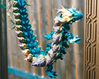May include: A colorful, articulated dragon sculpture in shades of blue, green, and yellow. The dragon is suspended in front of a wooden fence. The sculpture's design features a detailed, scaled body and spiky accents.